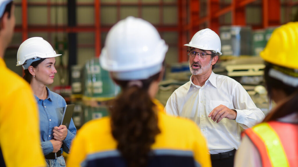 Professional Caucasian manager engineer wearing safety hardhat is training industry worker, workshop factory operator, mechanical engineering team production in heavy industrial manufacture factory.