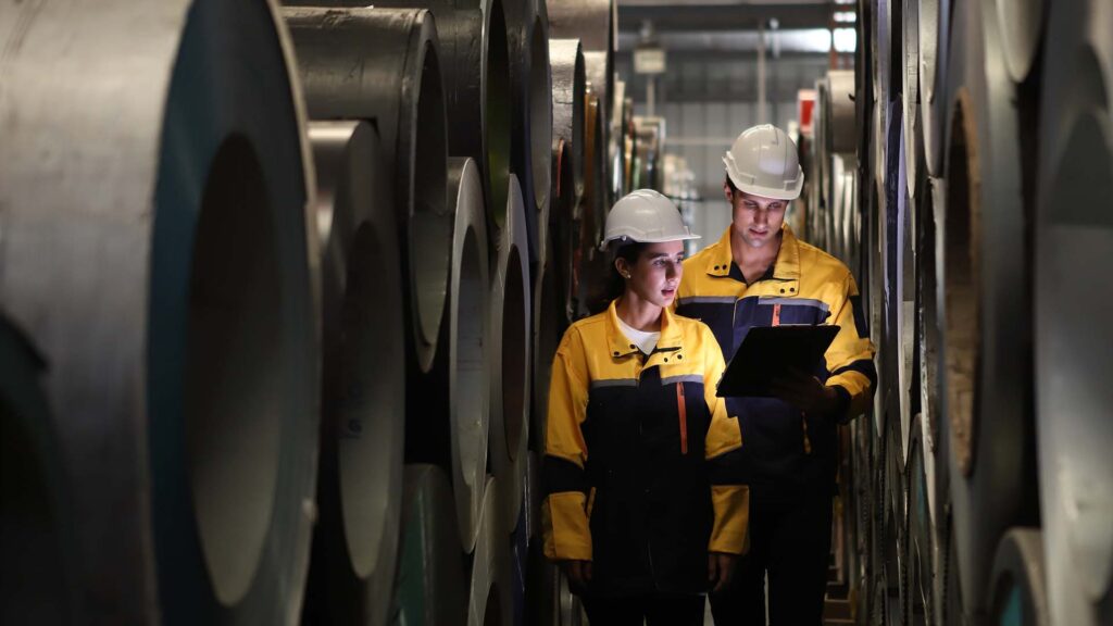 Workers in a factory surrounded by large metal tubes, with two workers passing in the middle
