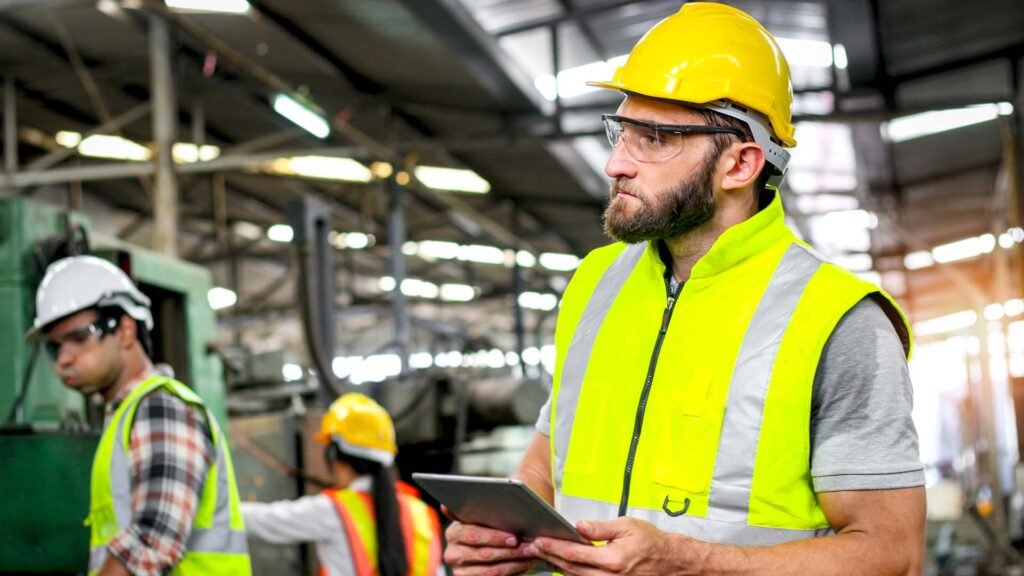 Industrial engineer with helmet and safety vest holds tablet for checking process and machinery machine engine at manufacturing factory. Worker man with safety glasses works in industry workplace.
