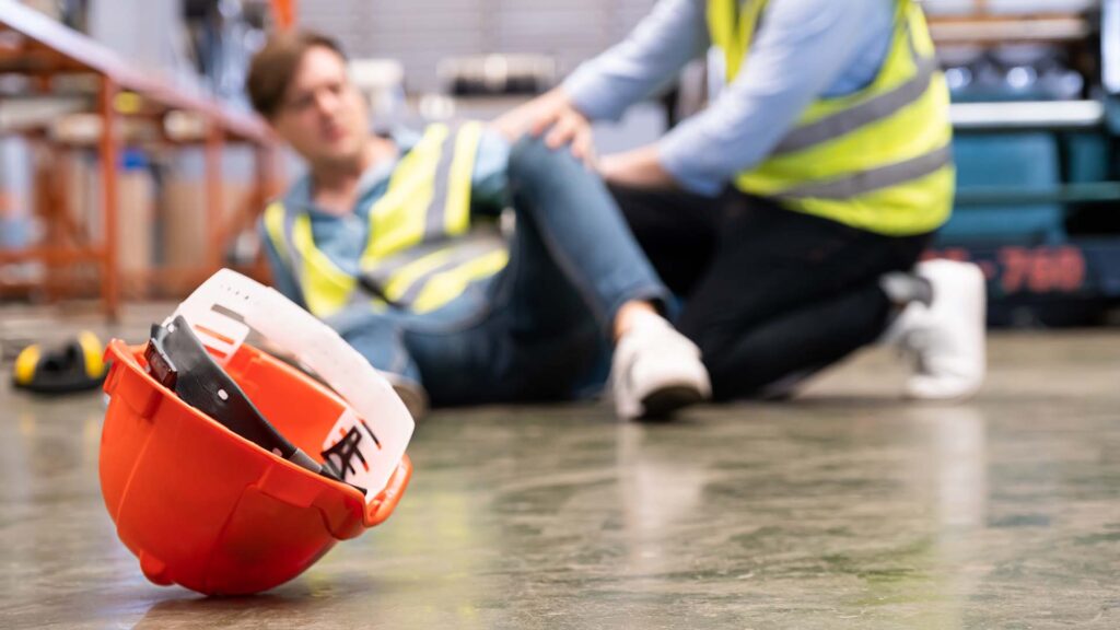 Close up of a construction worker's hard hat on the floor in focus with worker in the background on the floor hurt will co-worker is giving emergency assistance