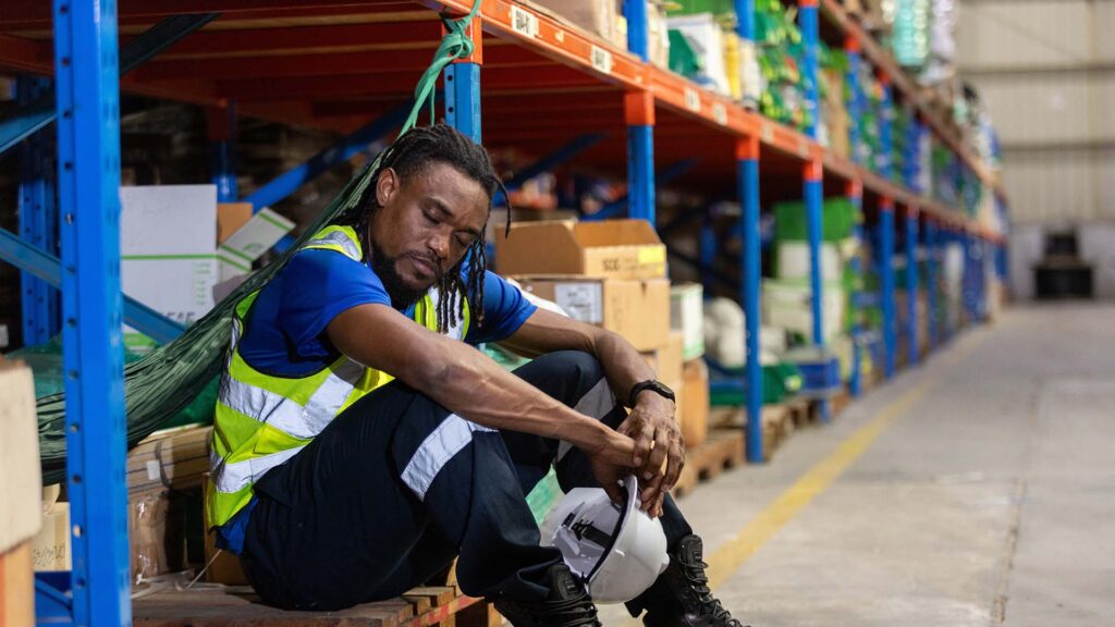 Exhausted warehouse worker in safety vest sitting on pallet with eyes closed, holding helmet, depicting fatigue, burnout, or emotional stress in logistics and industrial work environments.