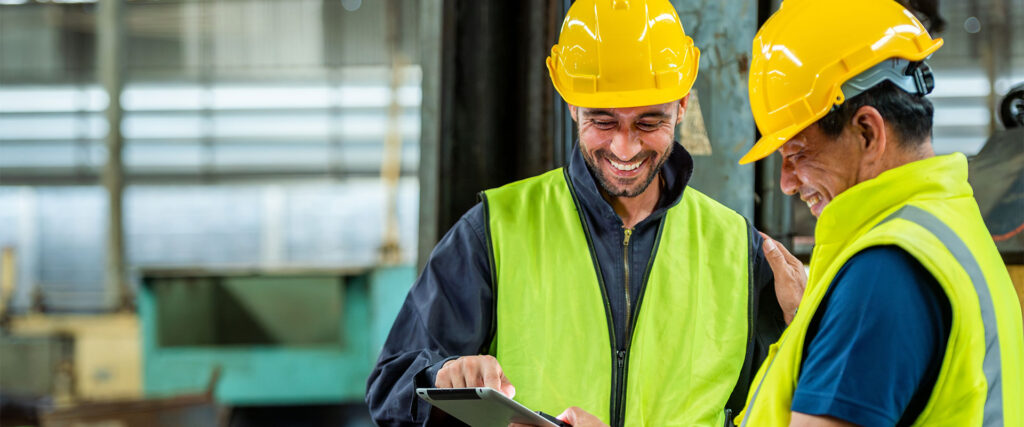 Two construction worker smiling reviewing documents on a tablet in a manufacturing plant